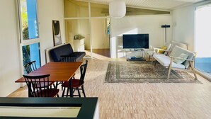 A modern living room with a wooden dining table, black and red chairs, a large flat-screen TV, and a white sofa with yellow cushions.
