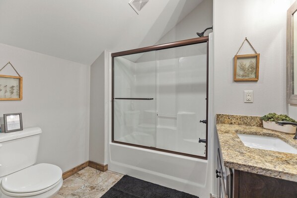 A bathroom with a glass-enclosed shower, a white toilet, a granite countertop, and a framed picture on the wall. - Lakefront, FirePit, Boat Slip & Golf Cart Included (Branson)