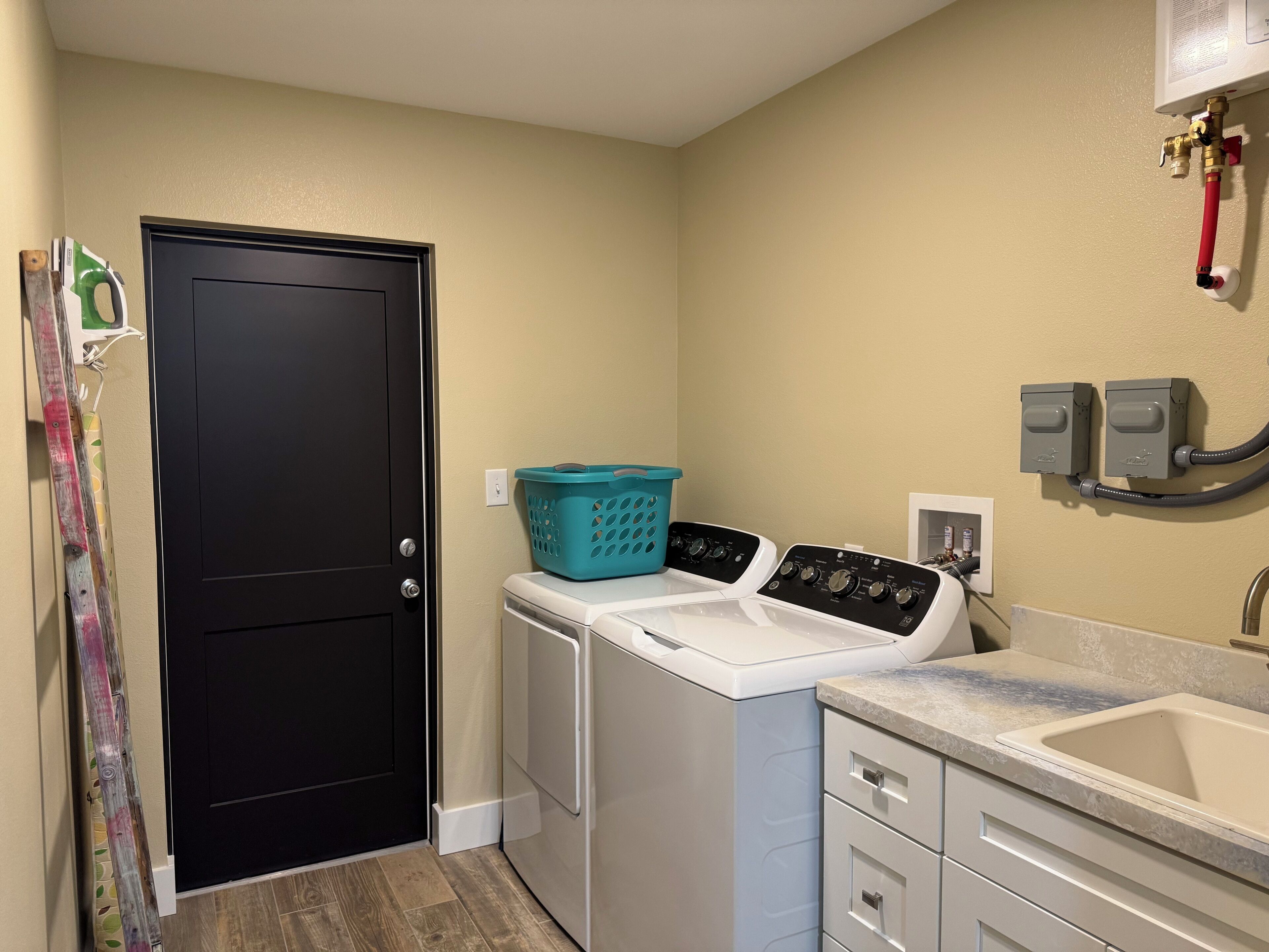 A laundry room with a black door, a washing machine, a dryer, a countertop with a sink, and two electrical outlets on the wall.