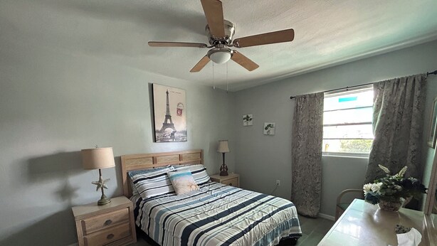 Ein Schlafzimmer mit Bett, Deckenventilator, Nachttisch, Lampe, Fenster mit Vorhängen und einem Wandbild.