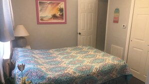 A bedroom with a patterned bedspread, a door, a wall-mounted picture, and a lamp.