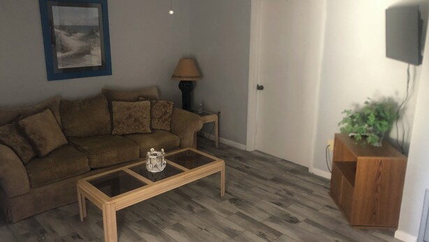 A living room with a brown sofa, a glass-top coffee table, a lamp, a door, and a television mounted on the wall.