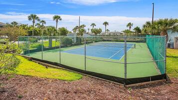 A tennis court surrounded by a chain-link fence, with palm trees and a clear sky in the background.