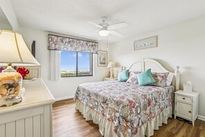 Un dormitorio con cama, mesita de noche, lámpara, ventilador de techo y ventana con vistas.