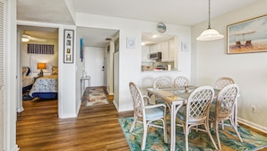 A dining area with a wooden table and wicker chairs, a kitchen with white cabinets, and a hallway leading to a bedroom with a bed and a nightstand. - Serene St. Augustine Ocean & Racquet 8315 Getaway (St. Augustine)