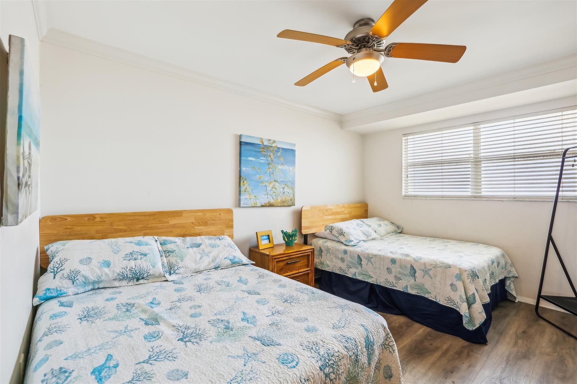 Une chambre avec deux lits, un ventilateur de plafond, une fenêtre avec des stores et un tableau au mur.