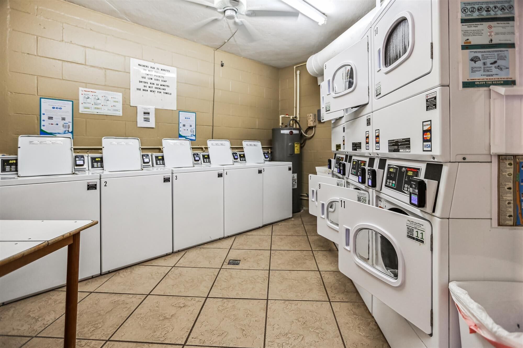 A laundry room with multiple washers and dryers, a ceiling fan, and informational posters on the walls.