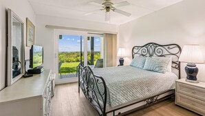A bedroom with a bed, a ceiling fan, a nightstand, a lamp, a dresser, a mirror, and a TV. - Oceanfront Luxury: 2BR Condo + Pool in St.aug (St. Augustine)