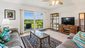 A living room with a ceiling fan, a television, a bookshelf, and a view through the glass door. - Oceanfront Luxury: 2BR Condo + Pool in St.aug (St. Augustine)