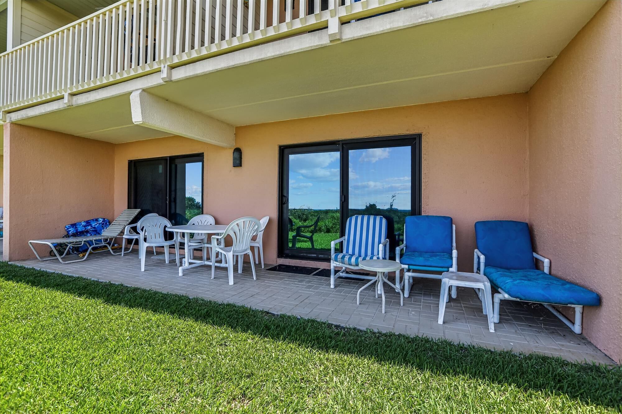 A balcony with white plastic chairs and a table, a blue lounge chair, and a view of a green field through large glass doors.