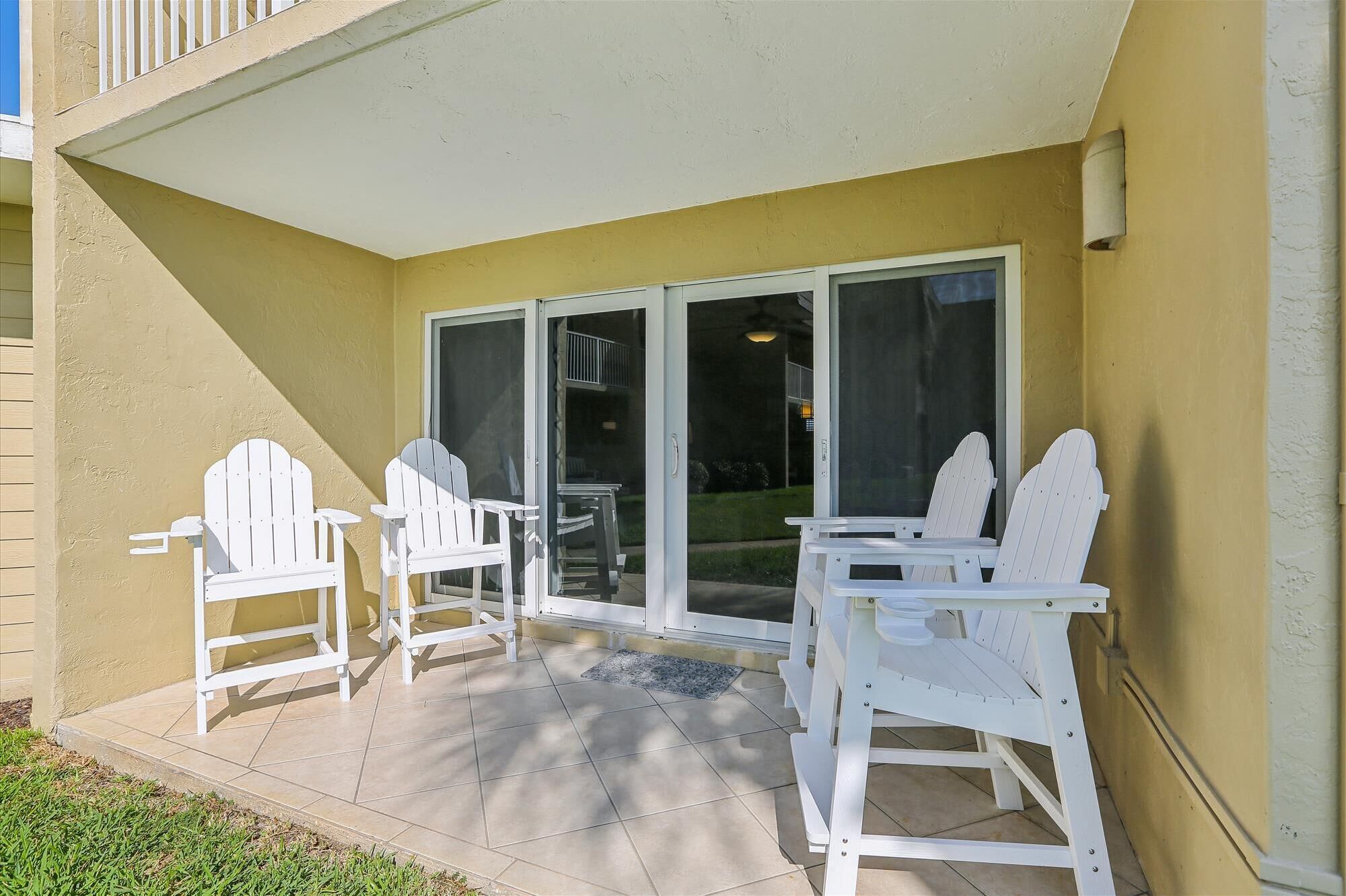 Un patio con dos sillas Adirondack blancas y una mesa, una puerta corredera de vidrio y una pared amarilla.