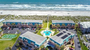 Un hotel frente a la playa con piscina central, varios estacionamientos y vista al mar.