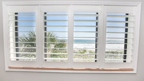 Una ventana con persianas horizontales, con vista a una playa y palmeras.