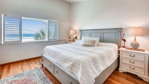 A bedroom with a bed, a nightstand, a lamp, a window with shutters, and a view of the ocean. - Coastal Oasis Refined Living Ocean Views (St. Augustine)