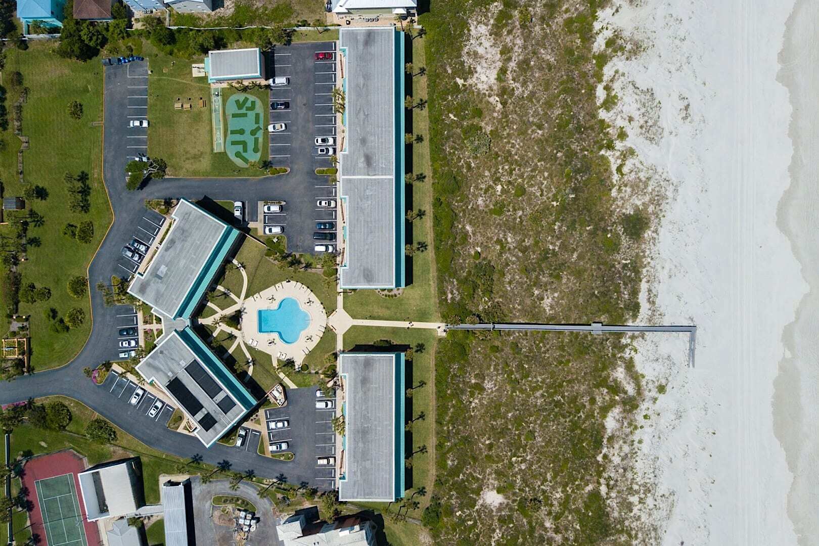 Vista aérea de um hotel à beira-mar, com piscina central, vários estacionamentos e um longo píer que se estende para o mar.