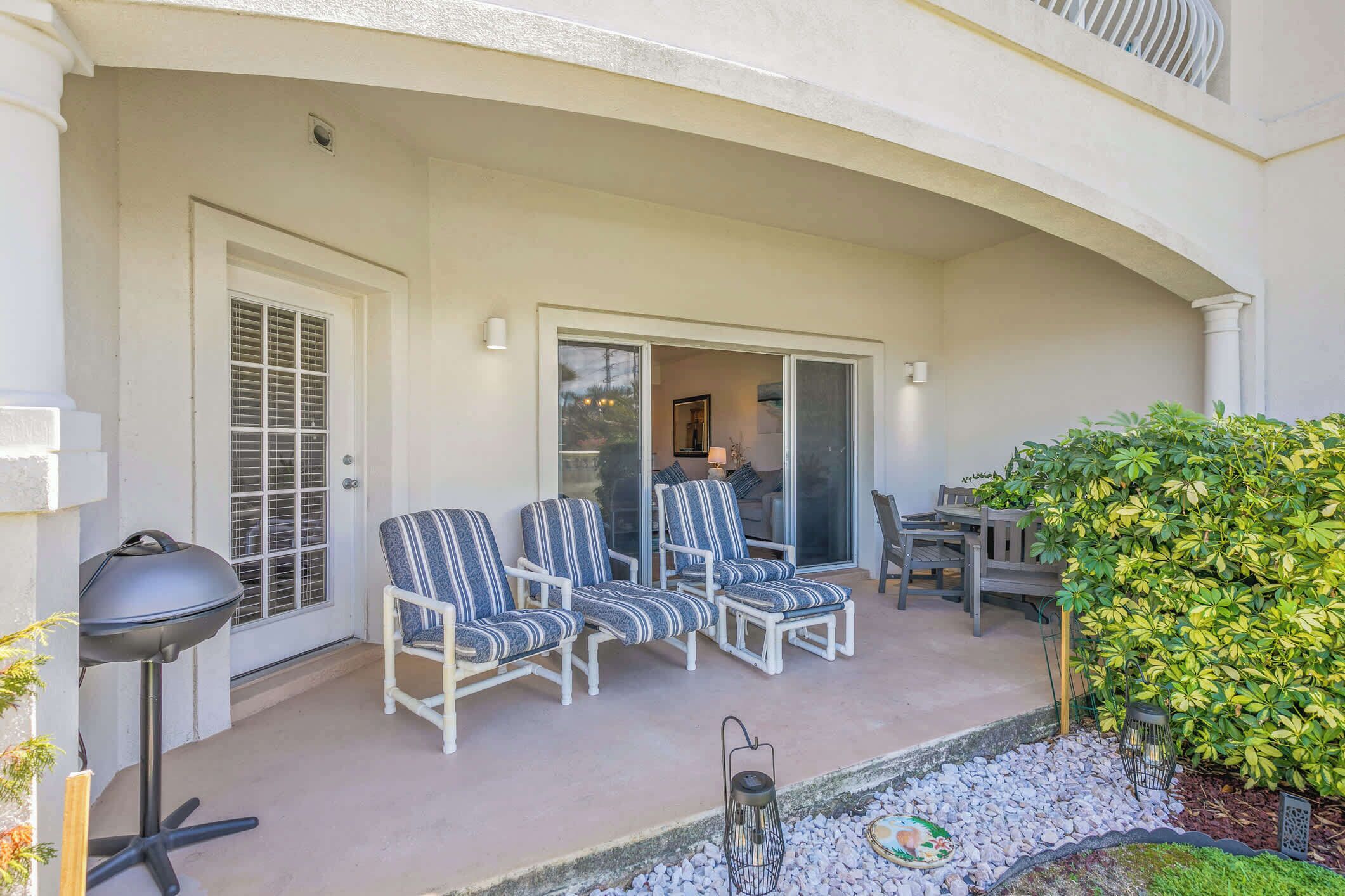 A covered outdoor seating area with white chairs and a table, a door with a window, and a small garden with a lantern.