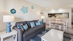 Una sala de estar con un sofá gris, cojines azules y blancos, una mesa de centro blanca, y una zona de comedor con una mesa y sillas blancas.