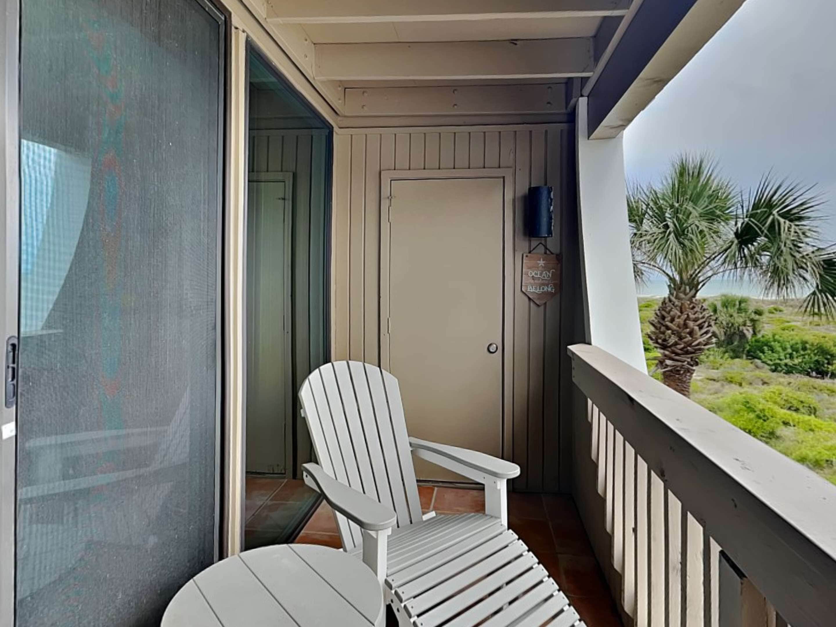 A balcony with a white Adirondack chair and a small table, a door, and a view of a palm tree and greenery.