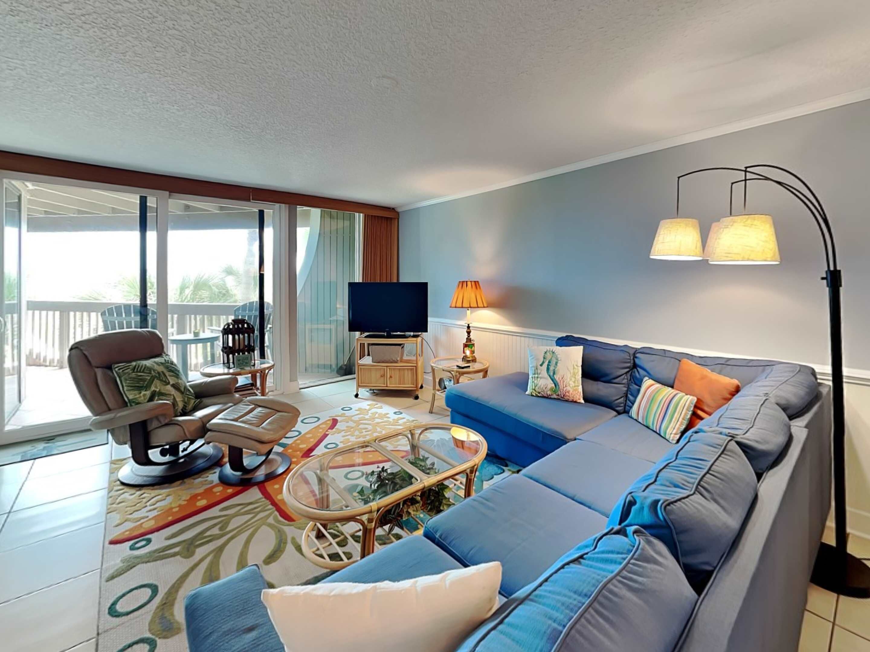 A spacious living room with a large sofa, a chair, a coffee table, and a TV. There is a balcony with a view of the outdoors.