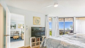 Ein Schlafzimmer mit einem großen Bett, einem Fernseher und einem Bücherregal. Es gibt einen Balkon mit Blick auf das Meer.