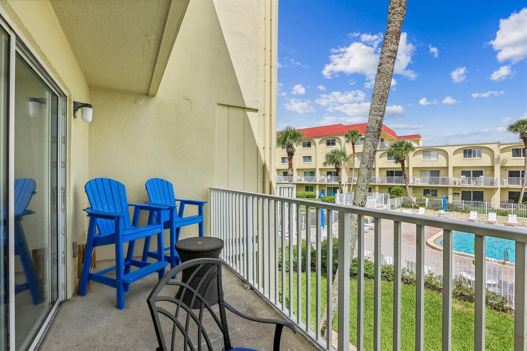 Un balcone con sedie blu, un tavolino e vista sulla piscina e sugli edifici dell'hotel.