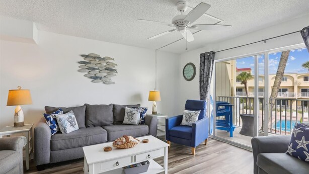 A modern living room with a grey sofa, blue armchair, and white coffee table. A ceiling fan and a wall clock are visible. - Beachfront Epic Ocean Views Sleeps 6 New Rental (St. Augustine)