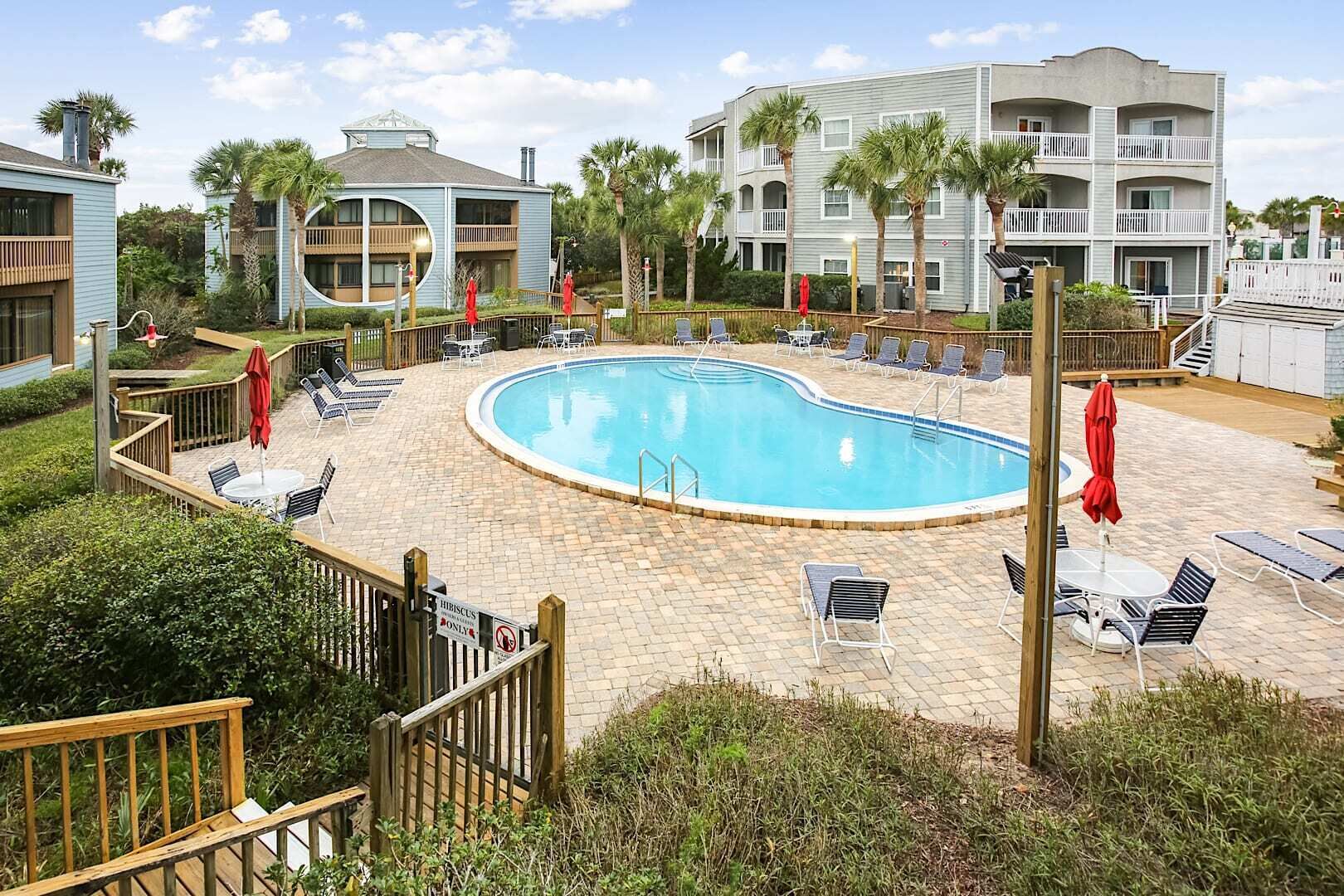 A swimming pool surrounded by lounge chairs and umbrellas, with a wooden deck and a view of a multi-story building.