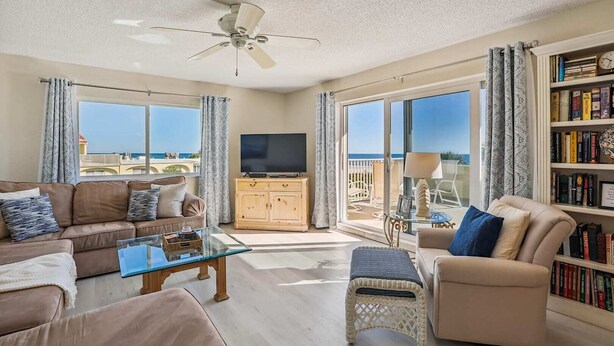 A spacious living room with a large flat-screen TV, comfortable seating, a bookshelf, and a balcony with a view of the sea. - Oceanfront 3BR Condo: Gated, Breathtaking View (St. Augustine)