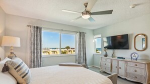 A hotel room with a large bed, a ceiling fan, a TV, a dresser, and a window with a view of the beach. - Oceanfront 3BR Condo: Gated, Breathtaking View (St. Augustine)