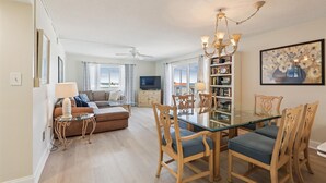 A spacious living room with a glass dining table, wooden chairs, a sofa, and a television. - Oceanfront 3BR Condo: Gated, Breathtaking View (St. Augustine)