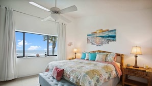 Een slaapkamer met een groot bed, een plafondventilator, een raam met uitzicht op het strand en twee nachtkastjes met lampen.