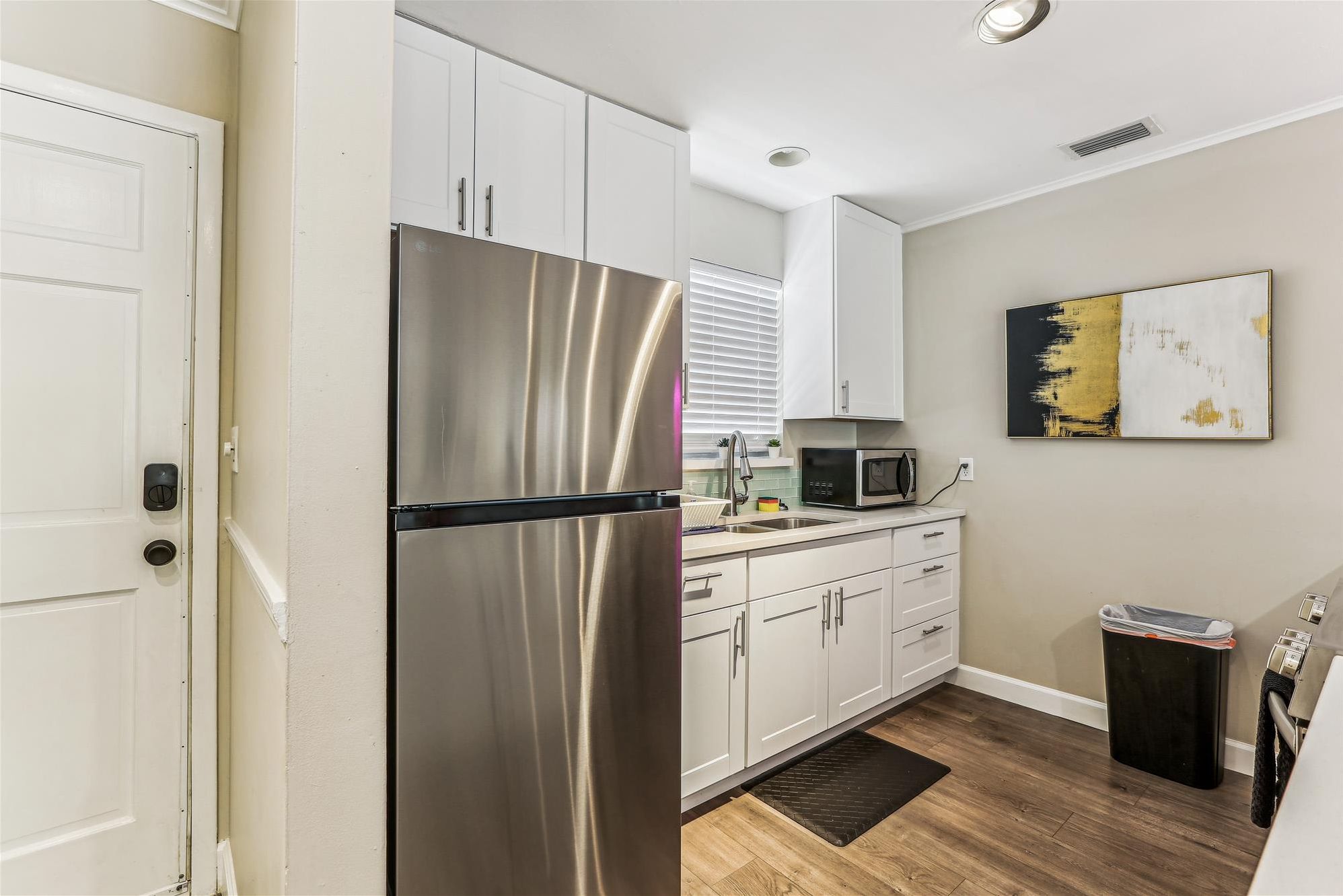 A compact kitchen with white cabinets, a stainless steel refrigerator, a microwave, and a black trash bin.