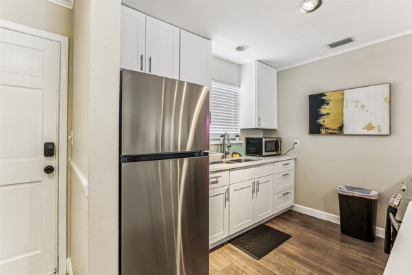 A compact kitchen with white cabinets, a stainless steel refrigerator, a microwave, and a black trash bin.