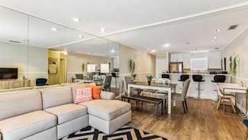 A modern living room with a sectional sofa, a dining area with a wooden table, and a kitchen with white cabinets.