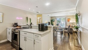 A modern kitchen with white cabinetry, a built-in oven, and a coffee maker. There is a dining area with a table and chairs, and a large window providing natural light.