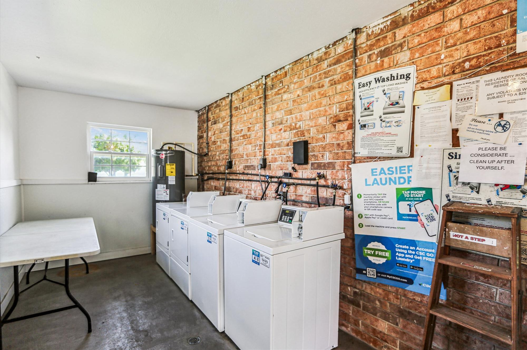 A laundry room with a brick wall, a water heater, and a washing machine. There are signs on the wall and a table with a chair.