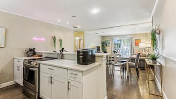 Een moderne keuken met witte kasten, een ingebouwde oven en een koffiezetapparaat. Er is een eethoek met een tafel en stoelen, en een groot raam zorgt voor natuurlijk licht.