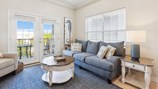 A living room with a grey sofa, a round coffee table, a side table, and a lamp. There is a door leading to an outdoor area with chairs.