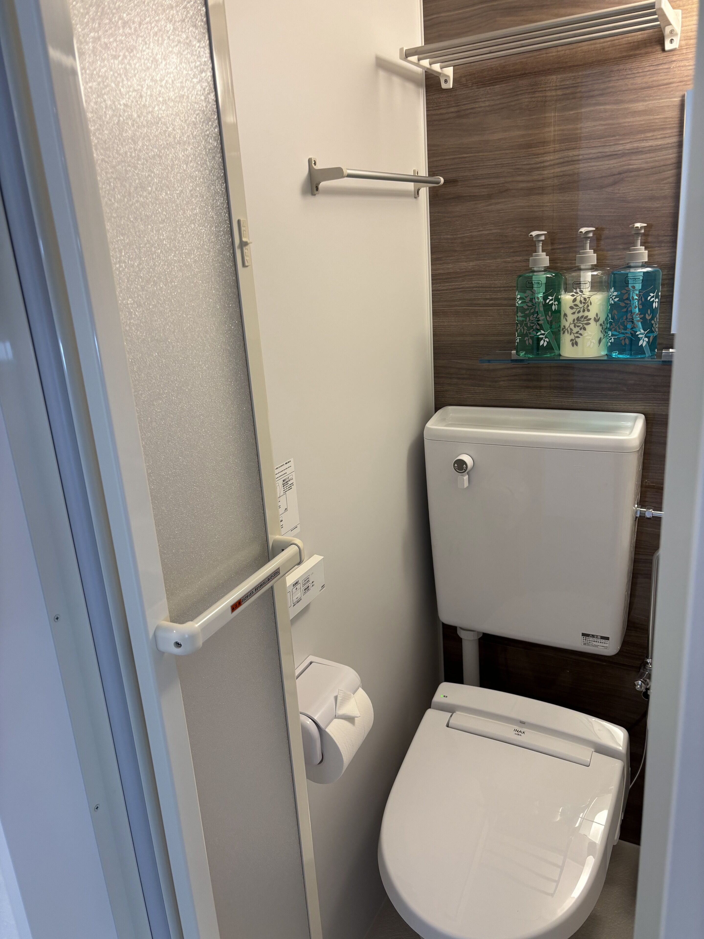Une petite salle de bain avec des toilettes blanches, une barre d’appui, une étagère pour le distributeur de savon et un porte-rouleau de papier toilette.