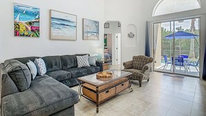 Ein Wohnzimmer mit einem dunkelgrauen Sofa, einem gemusterten Sessel und einem Couchtisch mit einem gewebten Korb. An der Wand hängen drei gerahmte Kunstwerke, ein Deckenventilator und ein Blick durch eine Glastür auf einen Außenbereich mit einem blauen Sonnenschirm.