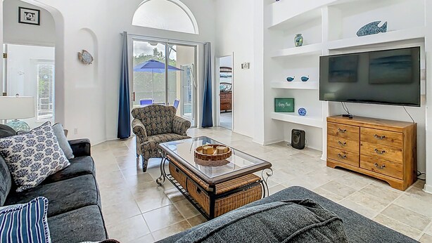 Ein geräumiges Wohnzimmer mit Sofa, Sessel und Couchtisch. Ein Fernseher steht auf einer Holzkommode, und ein großes Fenster ist mit Vorhängen versehen.