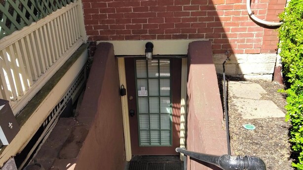 Un escalier extérieur mène à une porte surmontée d’une fenêtre, une boîte de raccordement est fixée au mur et une clôture verte se trouve à gauche.