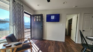 Una sala de estar con piso de madera, un televisor montado en la pared, una puerta y vista al exterior.