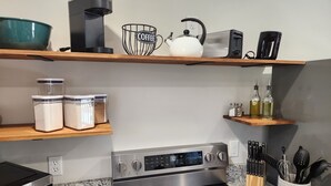 Una cocina con cafetera, hervidor, tostadora y diversos utensilios de cocina en estantes.