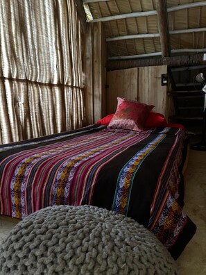A room with a bed, a red pillow, a striped blanket, and a knitted ottoman. - Ganesh Hostel (El Valle)