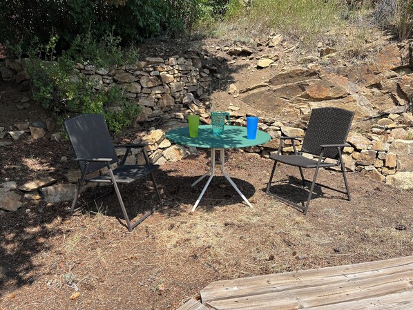Un área de estar al aire libre con dos sillas y una mesa, rodeada por un muro de piedra y vegetación natural.