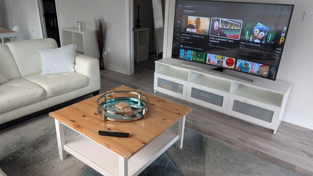 Un salon moderne avec un canapé blanc, une table basse en verre ornée de blocs de bois, une télécommande et un téléviseur à écran plat diffusant diverses vidéos.