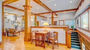 Un interior espacioso con vigas de madera, un comedor con mesa y sillas, una cocina al fondo y una escalera que lleva a un nivel superior.
