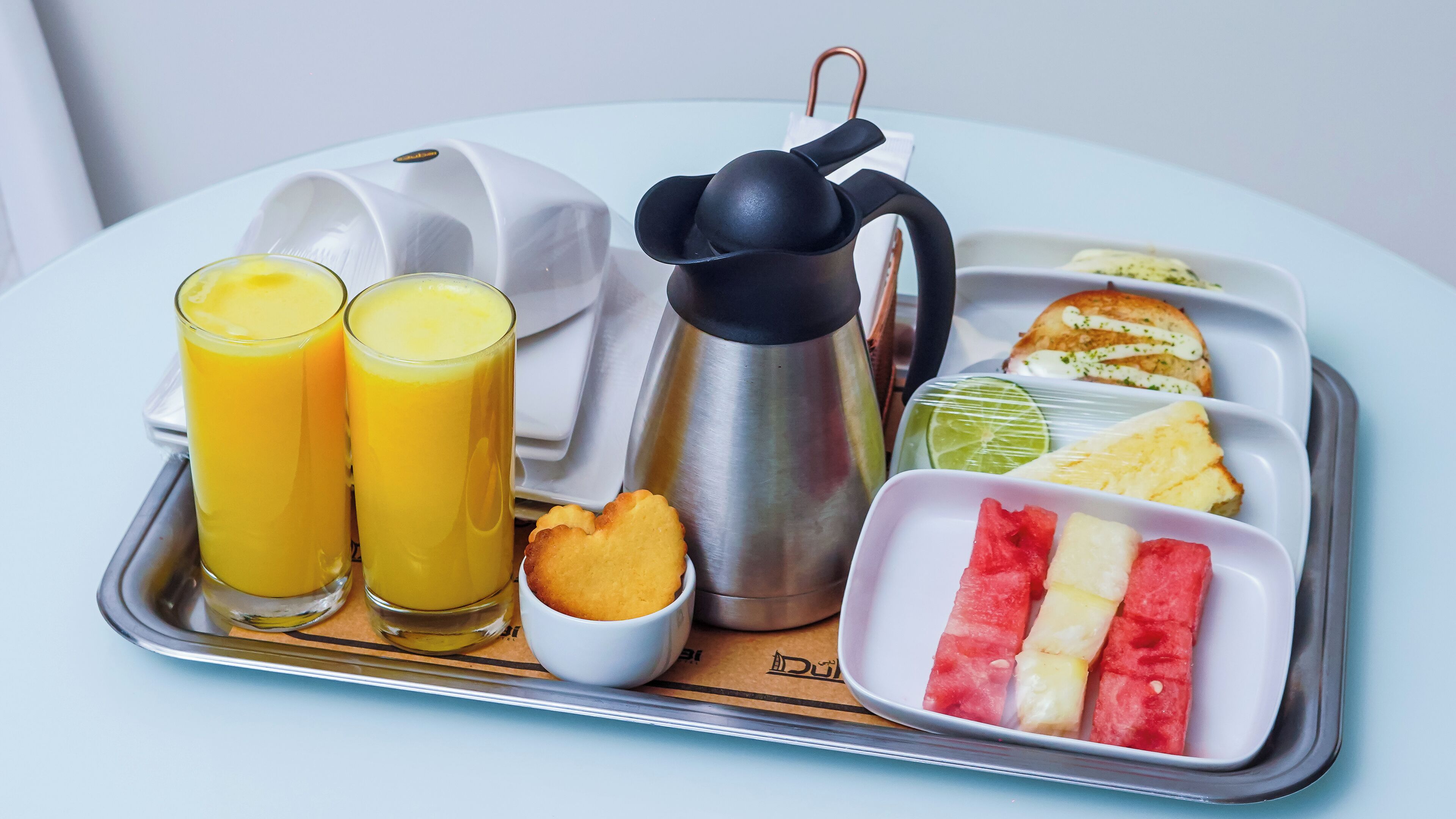 A tray with two glasses of orange juice, a thermos, a small bowl of cookies, a slice of toast with butter, a lime, a slice of pineapple, and a watermelon.