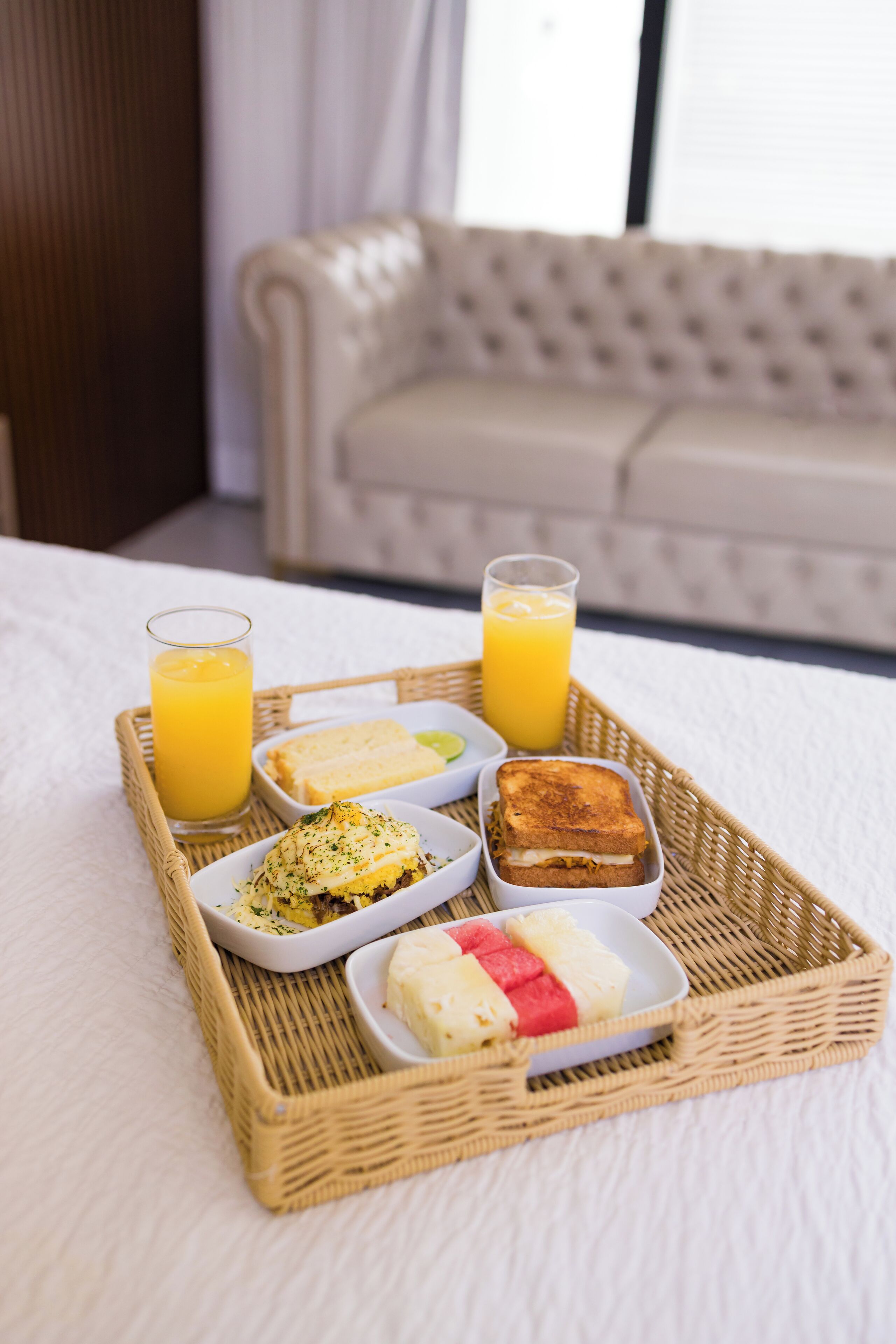 Un plateau de petit-déjeuner avec des toasts, des œufs brouillés, une salade de fruits et du jus d’orange, posé sur un couvre-lit blanc.